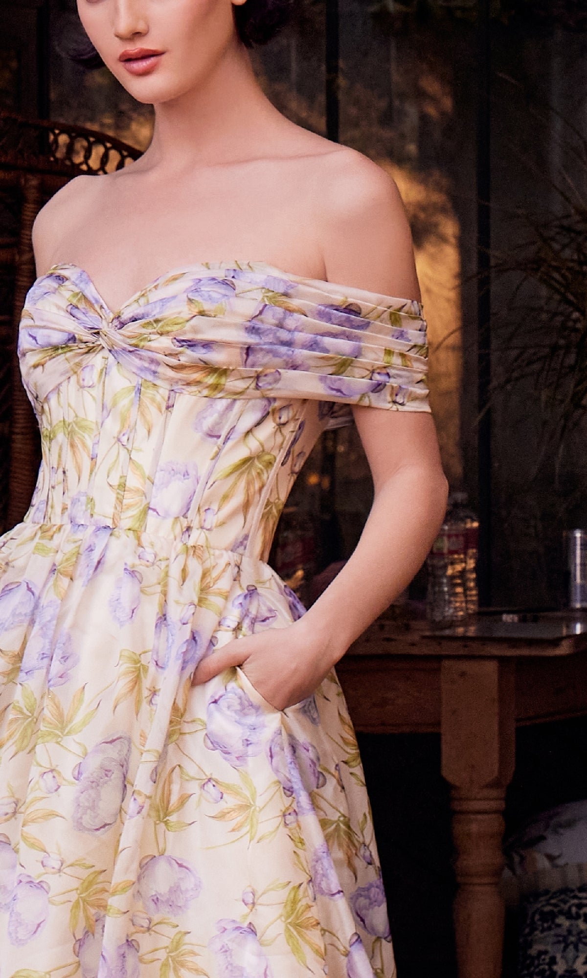 A woman stands indoors, partially out of frame, wearing the Andrea & Leo A1466 long floral-print prom dress featuring an off-shoulder design and pockets.